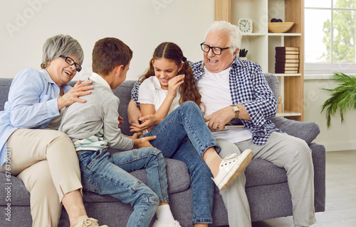 Happy grandparents and children having fun on the weekend. Funny, cheerful grandmother and grandfather tickling happy little kids grandchildren while sitting on the sofa at home all together photo