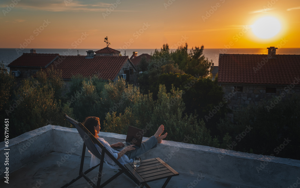 Happy young woman is working using laptop sitting on the patio in summer at sunset