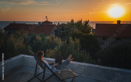 Obraz na płótnie Happy young woman is working using laptop sitting on the patio in summer at sunset w salonie