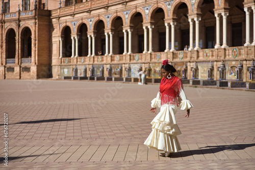 Young black and South American woman in a beige gypsy flamenco suit and red shawl, posing in a beautiful square in the city of Seville in Spain. Concept dance, folklore, flamenco, art.