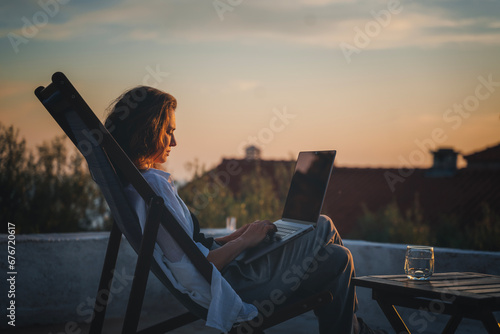 Happy young woman is working using laptop sitting on the patio in summer at sunset