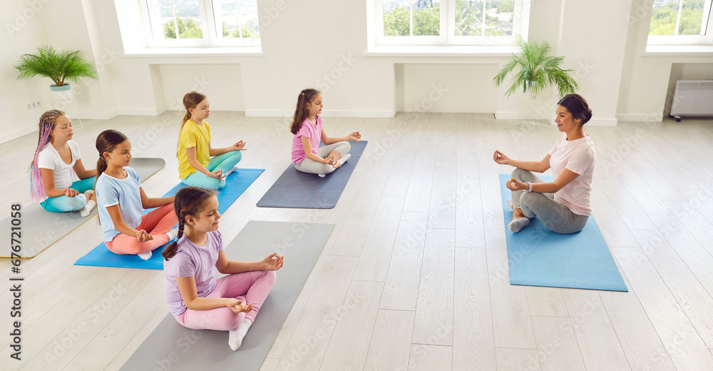 Side view portrait of kids girls sitting on the floor with female teacher trainer relaxing in gym sitting on yoga mat in Lotus pose meditating and doing yoga exercise. Children sport workout concept. - obrazy, fototapety, plakaty 