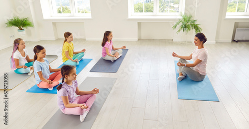 Side view portrait of kids girls sitting on the floor with female teacher trainer relaxing in gym sitting on yoga mat in Lotus pose meditating and doing yoga exercise. Children sport workout concept. photo