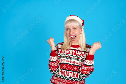 Excited young lady in festive holiday attire with Santa hat, jubilant against a vibrant blue backdrop.