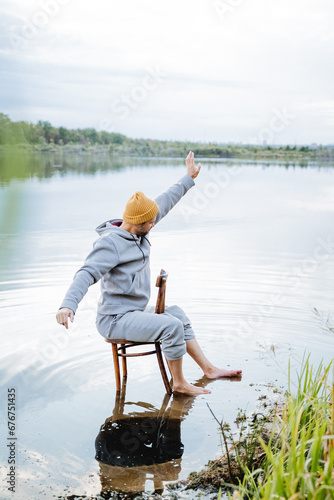 Man in water sitting on wooden chair, man stretching arms up, standing barefoot in cold pond.