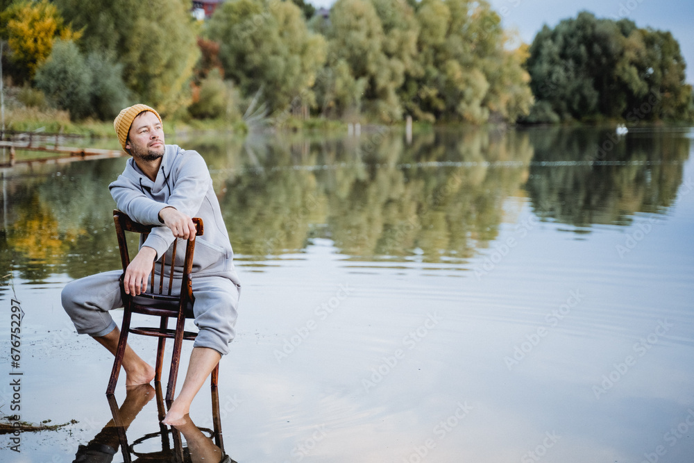 A guy on a chair is sitting in the water and looking away, morning photo shoot on the lake, a man is standing barefoot in the water.