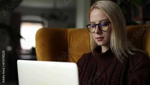 Caucasian woman in glasses and sweater with laptop sits in armchair at home