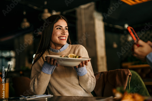 A smiling Latin woman smiling and holding a plate of food in the restaurant while the man is photographing her.