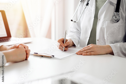 Doctor and patient sitting near each other at the white desk in clinic. Female physician is listening filling up a records form. Medicine concept