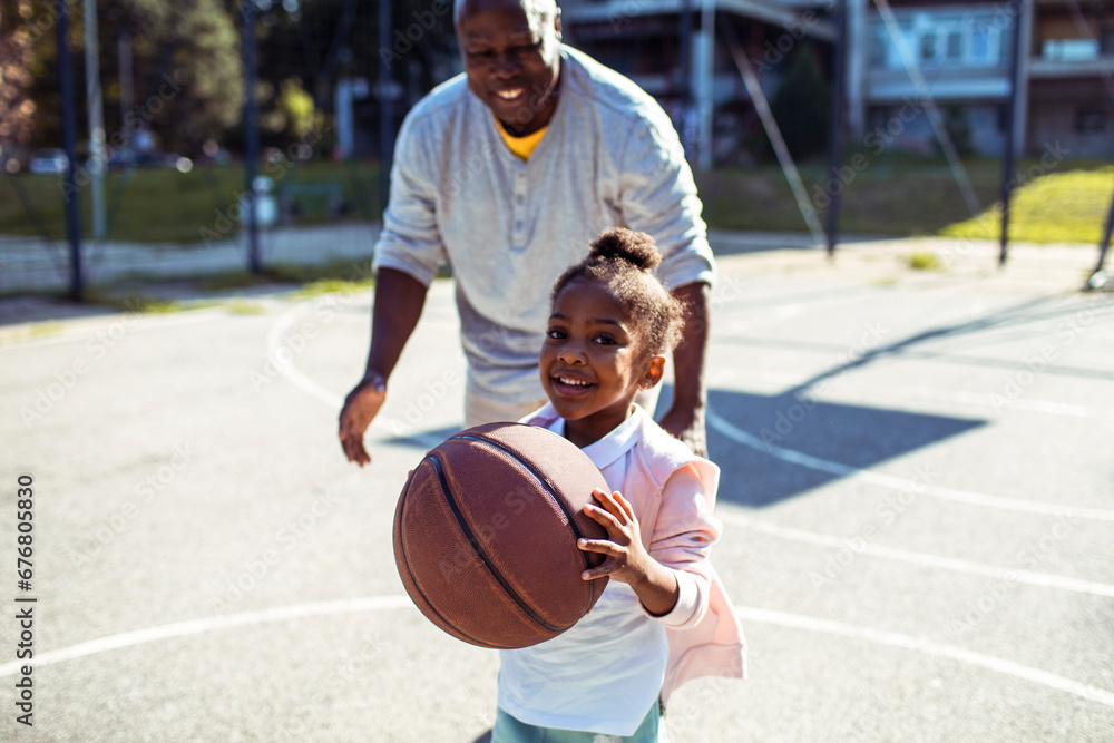 Adorable little girl playing basketball with father on outdoor court