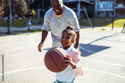 Adorable little girl playing basketball with father on outdoor court photo
