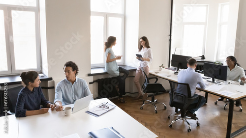 Multiethnic business team working in small office space, using laptop computer, cooperating on project, common tasks, talking at shared workplace tables. Modern rental co-working space