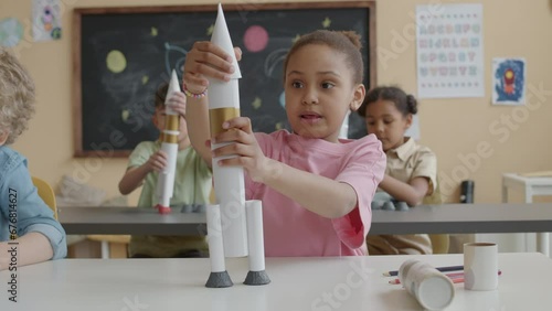 Little African American girl sitting at desk in primary school classroom, building paper rocket and raising it above head while playing during lesson photo