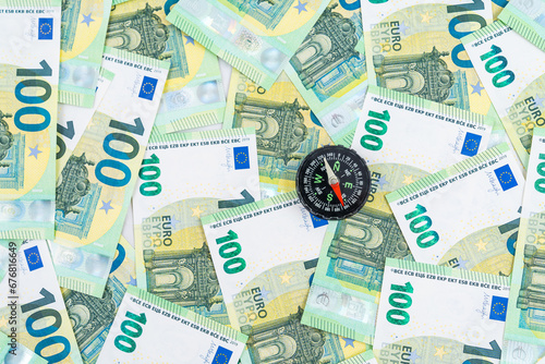  Top left view over a little black compass over many euro banknotes, 100 euro money bills, navigation in the world of finance concept