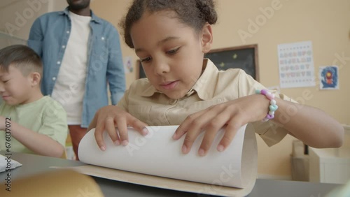 Little African American girl applying glue to paper and rolling it while making craft project on lesson at elementary schoolLittle African American girl applying glue to paper and rolling it while mak photo