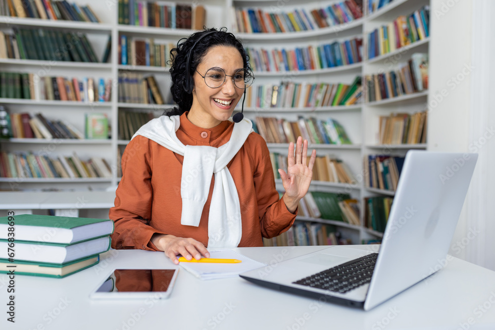 Joyful curly Hispanic girl sitting at a table in front of a laptop in a headset looking at the camera and waving at the camera, office worker in a call center, working video call. Joyful curly Hispanic girl sitting at a table in front of a laptop in a headset looking at the camera and waving at the camera, office worker in a call center, working video call.