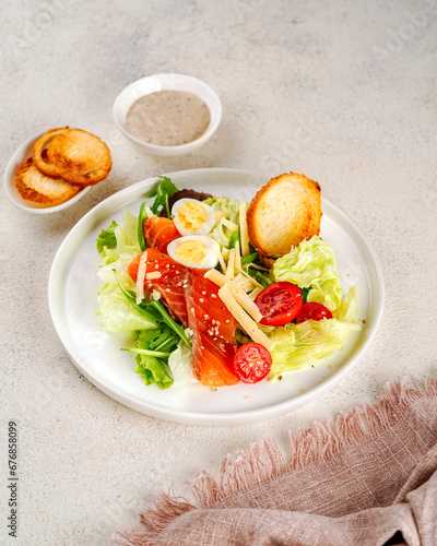 caesar salad with green lettuce, eggs, salmon, cherry tomatoes and croutons on a white ceramic plate, next to a bowl of sauce and a small plate of croutons
