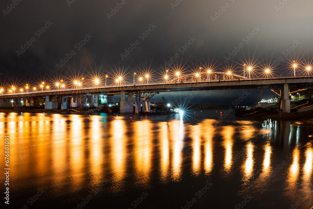 Bridge over the river illuminated by lanterns, reflection of lantern light on the water surface, road place night photo. Beautiful glare of lanterns.