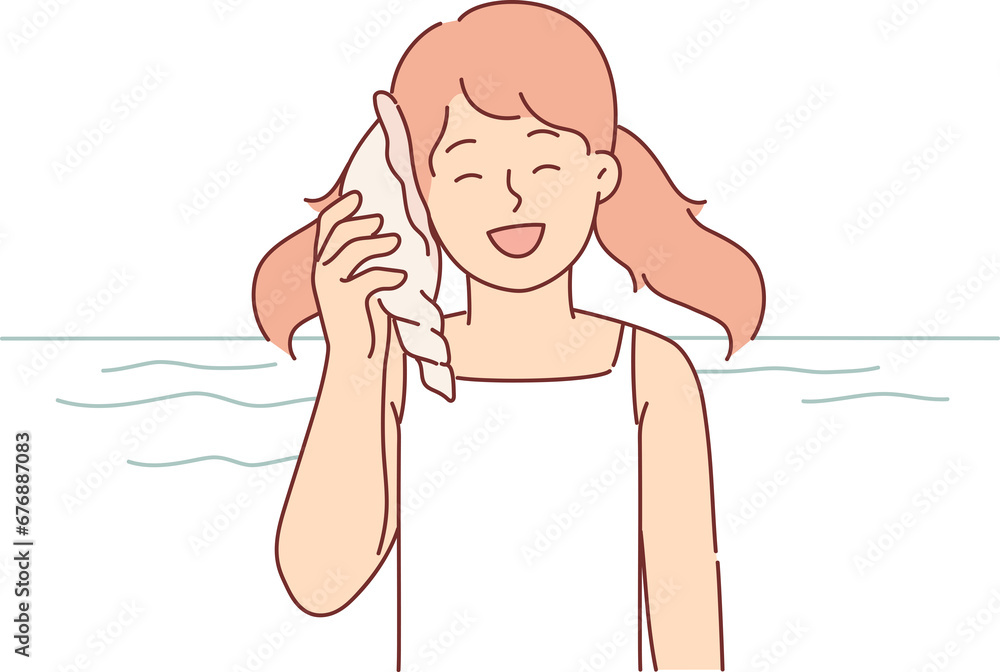 Little girl holds seashell to ear standing on beach of island, and smiles listening to sounds of sea Little girl holds seashell to ear standing on beach of island, and smiles listening to sounds of sea