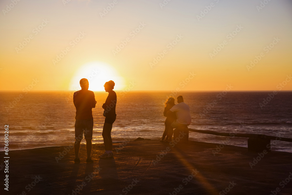 Silhouettes of the two pairs are on the pier against the background of bright sun and sunset sky. A group of people whose silhouettes are lit by the sunset on the horizon of the ocean.