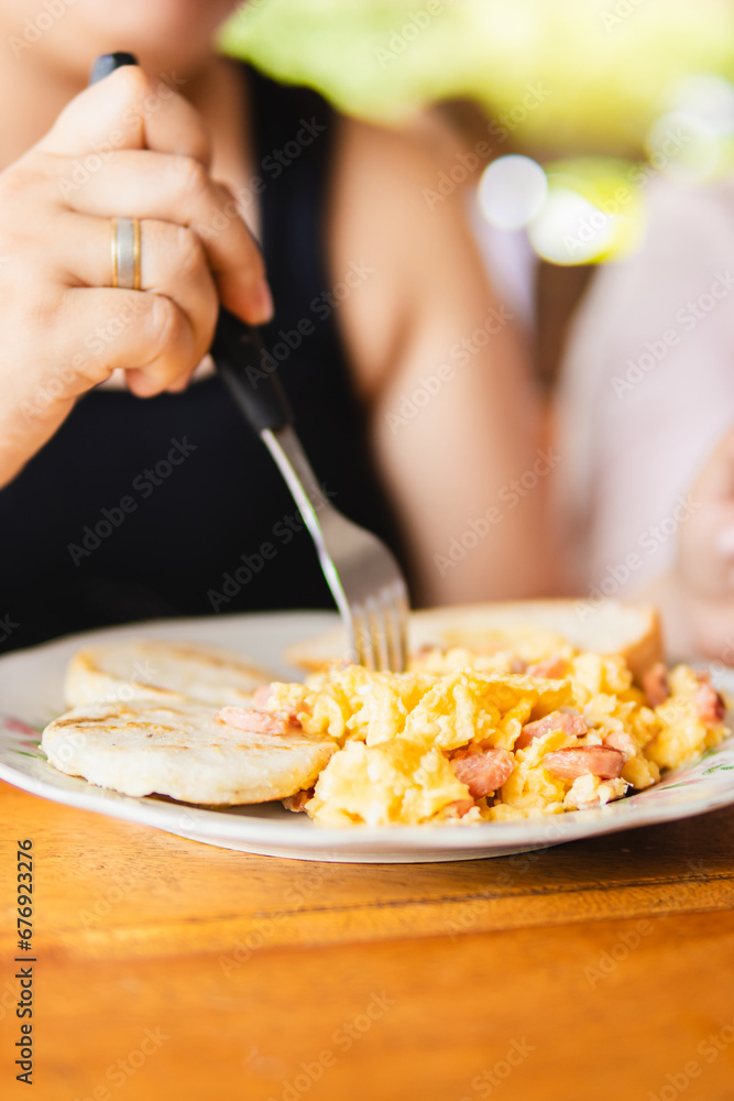Latina enjoying a typical Colombian breakfast with eggs, sausages, bread, and arepa, while sipping on chocolate and using a fork, promoting well-being for both body and mind Latina enjoying a typical Colombian breakfast with eggs, sausages, bread, and arepa, while sipping on chocolate and using a fork, promoting well-being for both body and mind