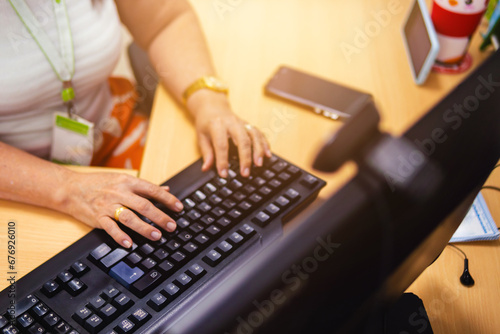 Senior woman working on a computer, her hands in the foreground; the PC is black in the office. There is a yellow glow, and a cell phone is placed next to the keyboard.