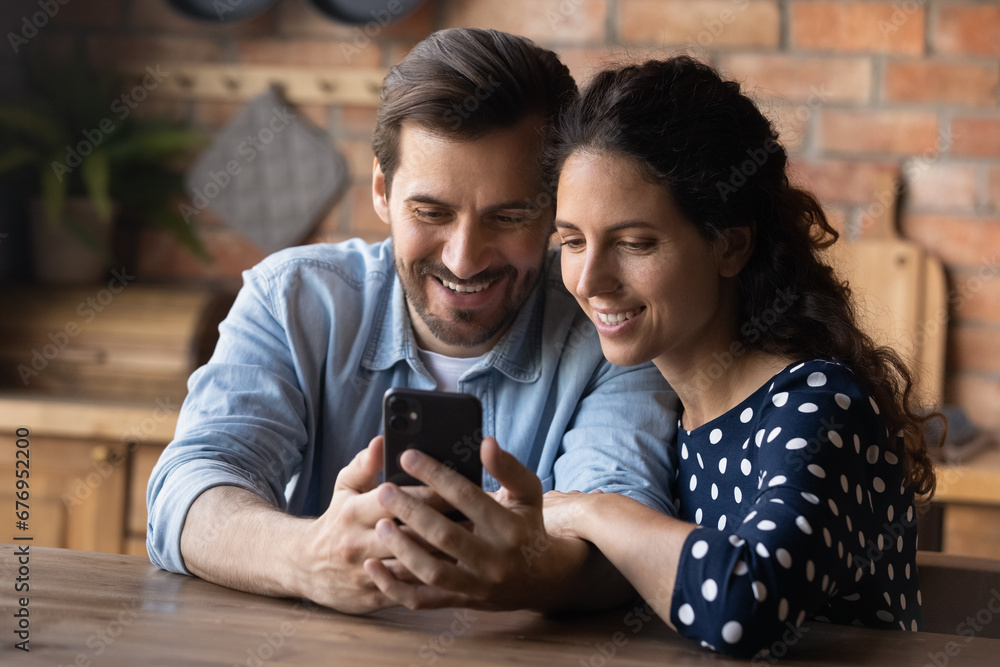 Close up smiling young couple hugging, using smartphone together, sitting at wooden table in kitchen, happy pleasant family enjoying leisure time, shopping or chatting in social networks online Close up smiling young couple hugging, using smartphone together, sitting at wooden table in kitchen, happy pleasant family enjoying leisure time, shopping or chatting in social networks online