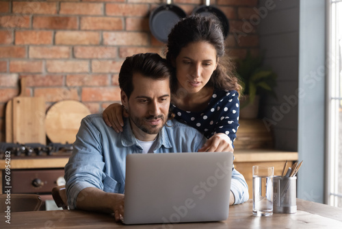 Serious young couple using laptop in cozy kitchen at home, focused woman and man looking at computer screen, chatting in social networks or shopping online, browsing banking service, surfing internet