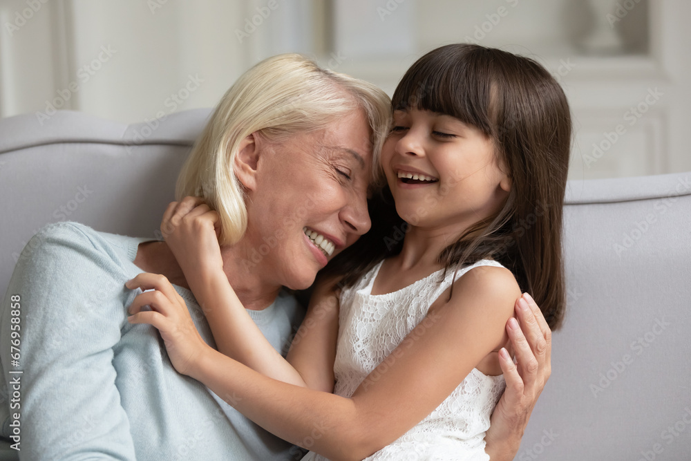 Happy mature grandmother and little granddaughter having fun, sitting on cozy sofa at home, adorable girl grandchild and smiling senior granny cuddling and laughing, spending leisure time together Happy mature grandmother and little granddaughter having fun, sitting on cozy sofa at home, adorable girl grandchild and smiling senior granny cuddling and laughing, spending leisure time together