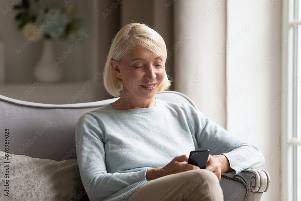 Smiling mature woman holding phone, sitting on cozy sofa at home, happy older female looking at smartphone screen, shopping or chatting online, reading message, watching video in social network Smiling mature woman holding phone, sitting on cozy sofa at home, happy older female looking at smartphone screen, shopping or chatting online, reading message, watching video in social network