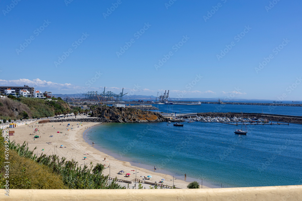 View to sines beach with industrial container port terminal cranes behind, in the city of Sines on the South of Portugal