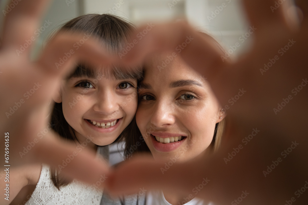 Head shot portrait close up funny cute little girl and young mother showing heart gesture with hands, smiling daughter and parent looking at camera through fingers, child and mum connection and unity Head shot portrait close up funny cute little girl and young mother showing heart gesture with hands, smiling daughter and parent looking at camera through fingers, child and mum connection and unity