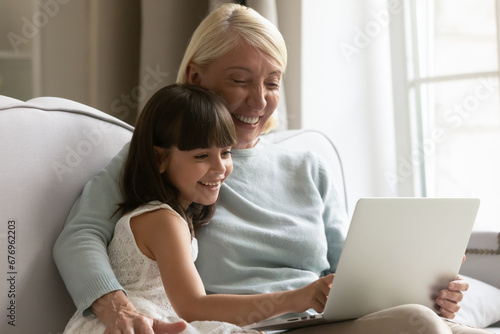 Happy mature grandmother and cute little granddaughter having fun with laptop together, looking at screen, sitting on sofa at home, smiling girl grandchild and senior granny using computer close up