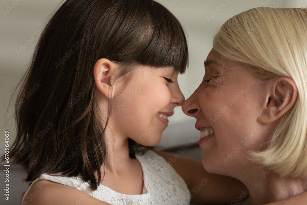 Close up happy mature grandmother and cute little girl touching noses, enjoying tender moment, adorable granddaughter and smiling senior grandma cuddling, expressing love, two generations Close up happy mature grandmother and cute little girl touching noses, enjoying tender moment, adorable granddaughter and smiling senior grandma cuddling, expressing love, two generations