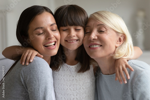 Head shot portrait three generations of women hugging, smiling mature grandmother, young mother and little preschool daughter cuddling with closed eyes, granny, mum and granddaughter embracing