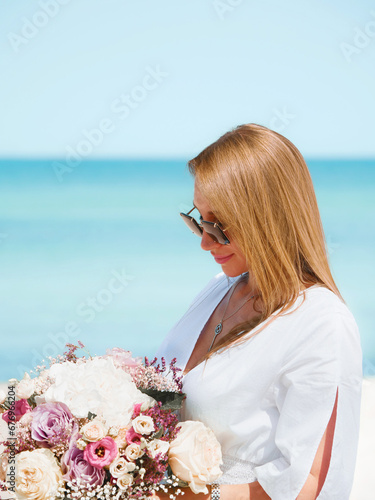 Obraz na płótnie Joyful Young Woman Holding a Bouquet of Flowers on a Beach with Blue Sky Background w salonie