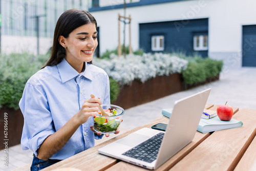 Happy woman holding fresh salad while working on laptop sitting outside building office  looking at screen  watching video or listening colleague on video call. Business concept.