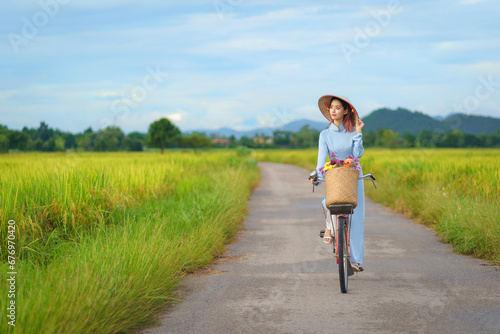 Beautiful Vietnamese Asian woman wearing a blue color Ao Dai National Costume Dress with red bicycle and flowers fresh yellow rice fields mountain background. Portrait fashion show in nature.