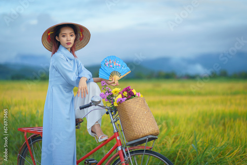 Beautiful Vietnamese Asian woman wearing a blue color Ao Dai National Costume Dress with red bicycle and flowers fresh yellow rice fields mountain background. Portrait fashion show in nature.