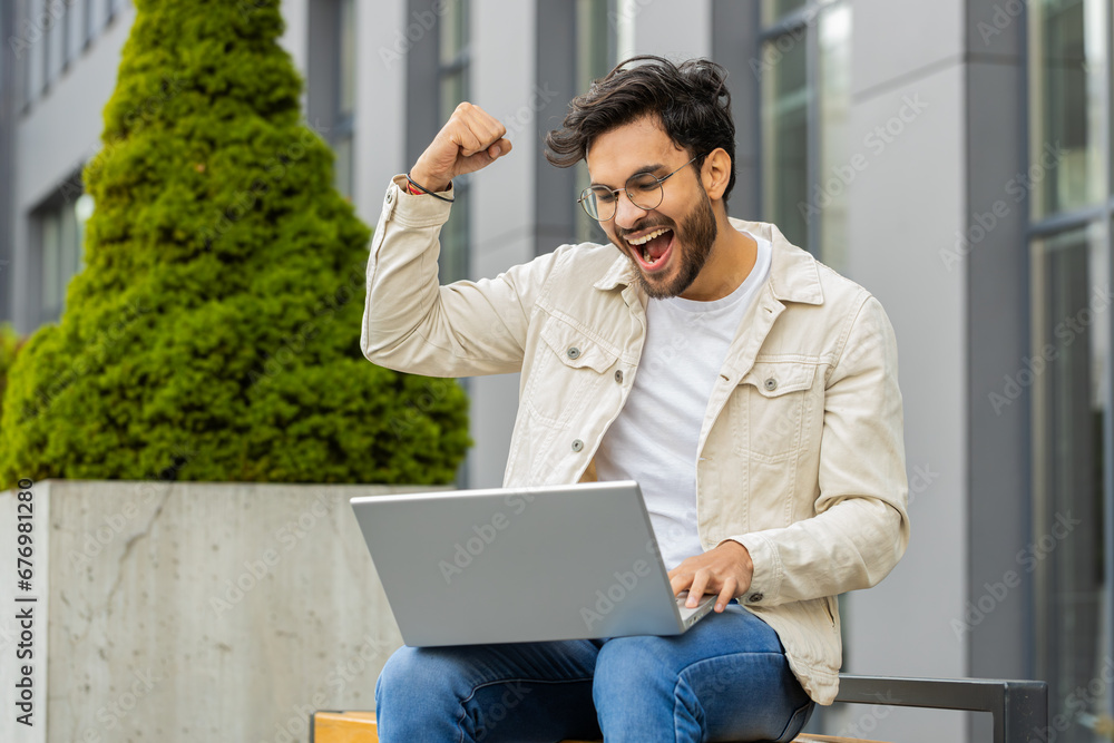 Overjoyed happy young Indian man working on laptop scream in delight raise hands in triumph winner gesture celebrate success win money outdoors. Arabian Hindu guy sitting on downtown street bench Overjoyed happy young Indian man working on laptop scream in delight raise hands in triumph winner gesture celebrate success win money outdoors. Arabian Hindu guy sitting on downtown street bench