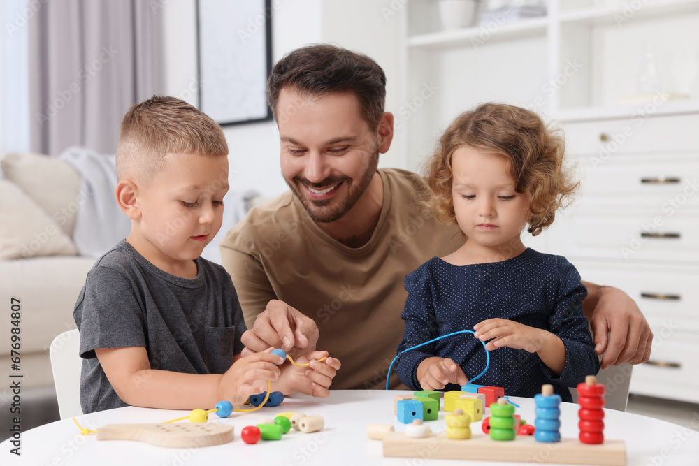 Motor skills development. Father and his kids playing with wooden pieces and string for threading activity at table indoors Motor skills development. Father and his kids playing with wooden pieces and string for threading activity at table indoors