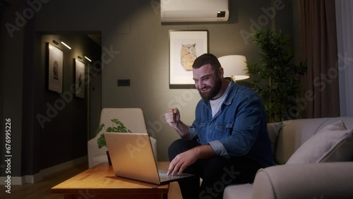 Bearded young adult man sitting on sofa while using laptop checking messages read notice receive good news on e-mail celebrate great opportunity feels happy. Special offer, student got scholarship photo