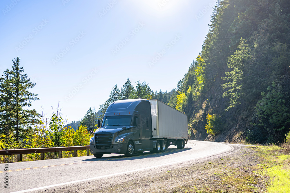Stylish dark gray big rig semi truck transporting cargo in dry van semi trailer running on the winding mountain road