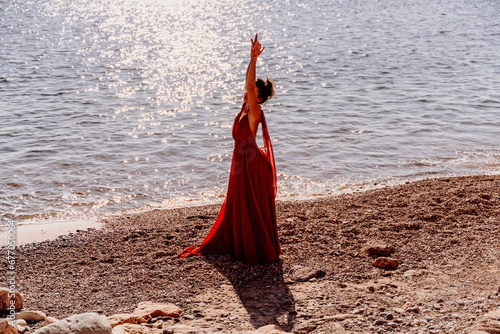 Woman red dress sea. Woman in a long red dress posing on a beach with rocks on sunny day. Girl on the nature on blue sky background.