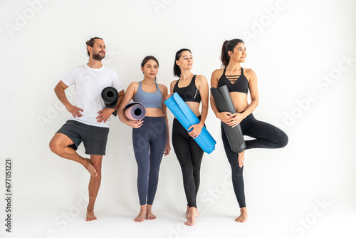 Group of young sporty girl, three friends and one man with yoga mats standing at white wall. Female companions in gym resting after fitness, indoor full length in loft studio.Healthy lifestyle concept