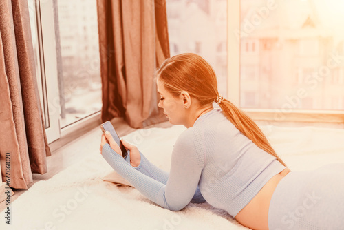 Side view portrait of relaxed woman with long hair lying on carpet at home. She is dressed in a blue tracksuit, holding a phone in her hands.