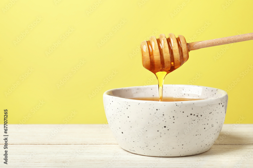 Pouring tasty honey from dipper into bowl at white wooden table against yellow background, closeup. Space for text Pouring tasty honey from dipper into bowl at white wooden table against yellow background, closeup. Space for text