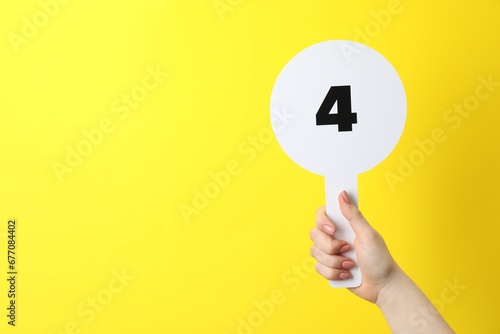 Woman holding auction paddle with number 4 on yellow background, closeup. Space for text