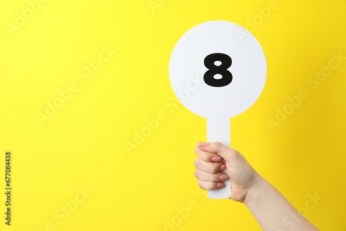 Woman holding auction paddle with number 8 on yellow background, closeup. Space for text