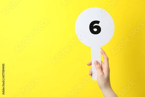 Картината върху платно Woman holding auction paddle with number 6 on yellow background, closeup